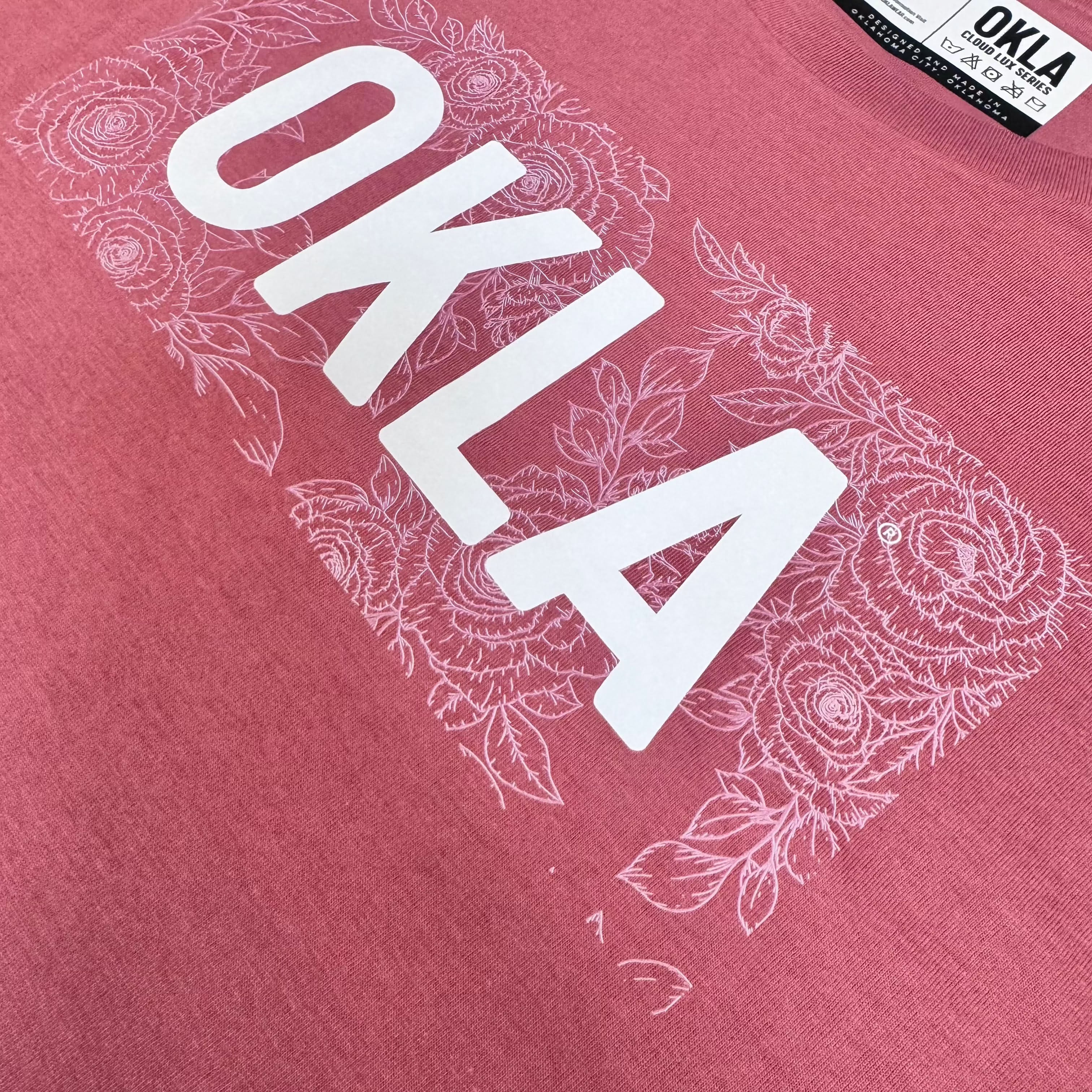 The stylish OKLA Rose Crop Top Tee, made from breathable material, features the word OKLA in striking white letters amidst detailed floral patterns. With an OKLA branding tag at the collar, this pink t-shirt seamlessly combines comfort and fashion.