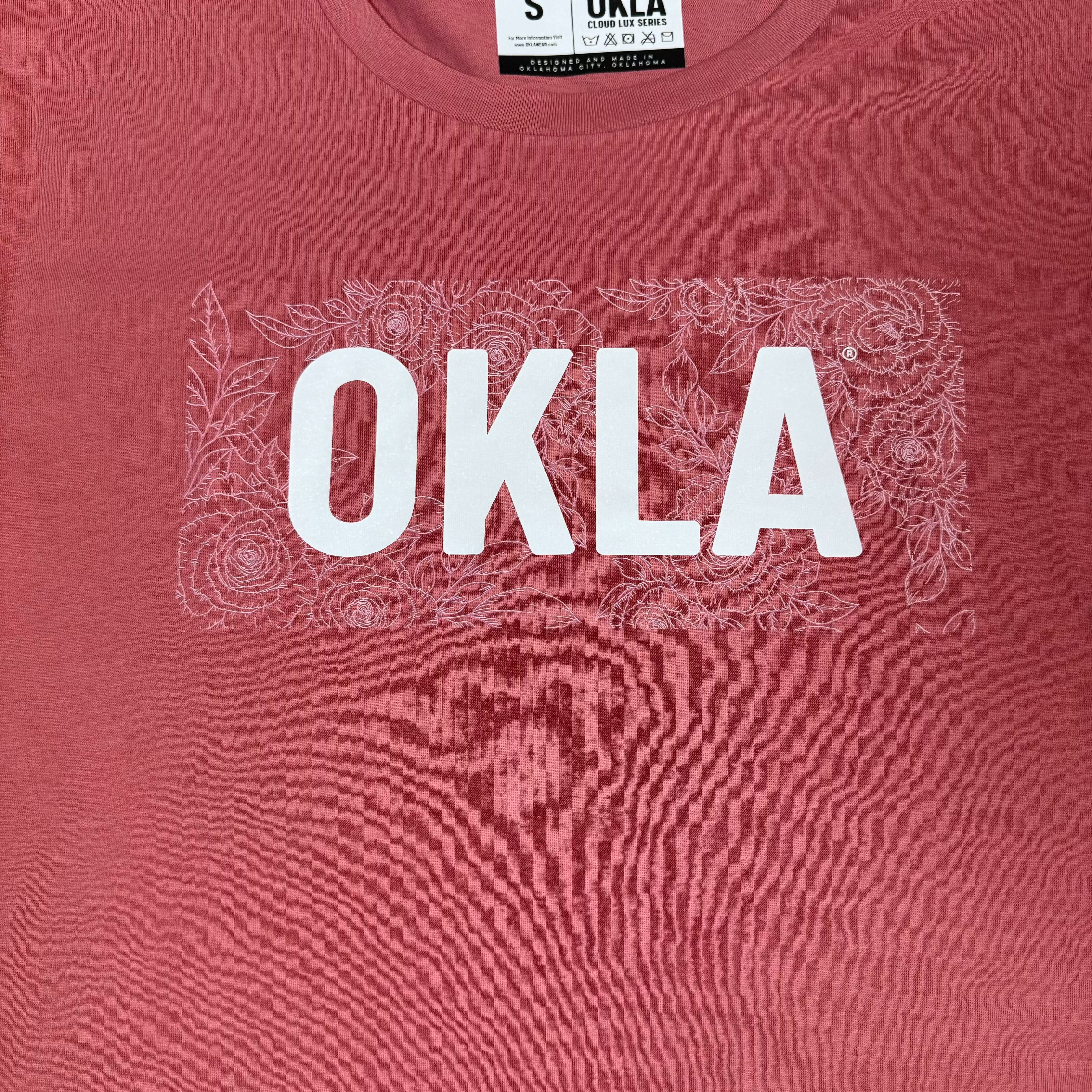The OKLA Rose Crop Top Tee showcases a striking red design with the word "OKLA" boldly displayed in white letters across the chest, complemented by a subtle floral pattern. Made from breathable fabric, this chic crop top ensures both comfort and style.