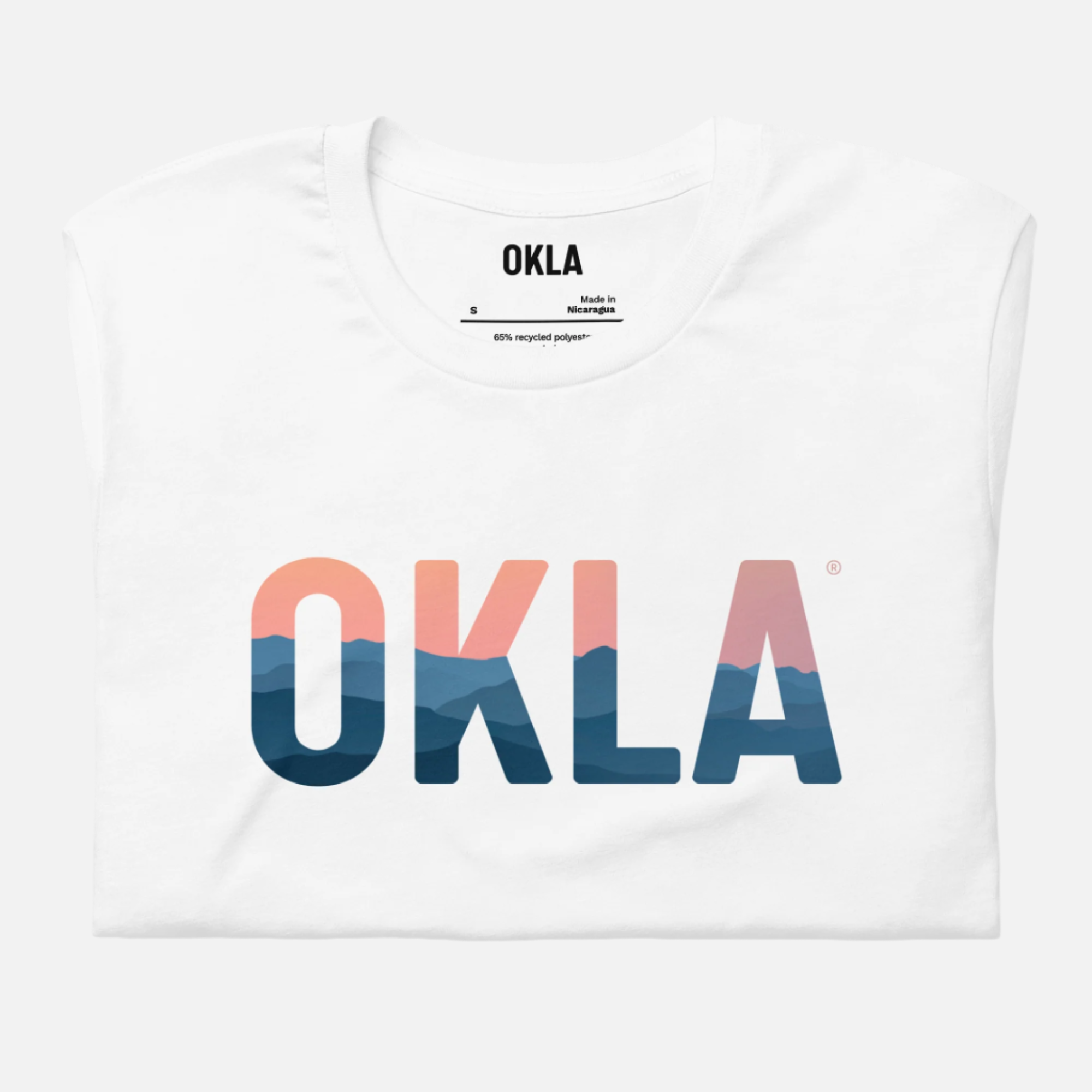 Introducing the OKLA Horizon Tee: a folded white T-shirt featuring "OKLA" printed with striking blue and pink mountain silhouettes. Made from recycled cotton, it ensures a blend of adventure-ready comfort and sustainability. The label underscores OKLA's dedication to quality and eco-friendly materials.