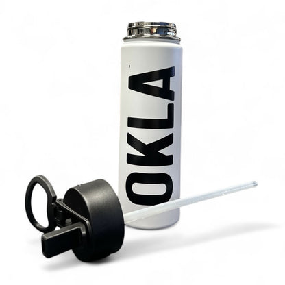 Introducing the OKLA - Travel Thermos 22oz: This sleek white stainless steel water bottle is adorned with the bold black letters "OKLA" printed vertically. It comes with a leak-proof lid, complete with an attached black plastic handle and straw, making it an ideal travel companion. The minimalist design features a plain white background, enhancing its stylish appearance.