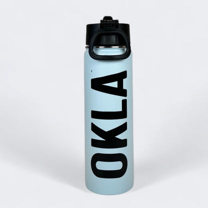 A light blue OKLA - Travel Thermos 22oz stands upright, featuring a leak-proof black lid. Crafted from stainless steel, the thermos showcases bold black letters spelling OKLA vertically down the front, set against a plain white background.