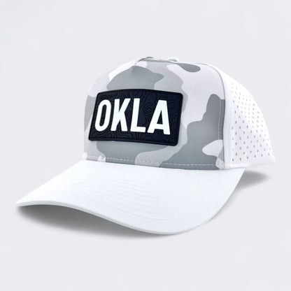 The OKLA Navigator Hat, in Snow Camo, is a baseball cap featuring sweat-wicking technology and a perforated back panel. Its front displays a vintage-inspired 3D patch with 'OKLA' in bold white letters on a black background, set against a simple light gray backdrop.