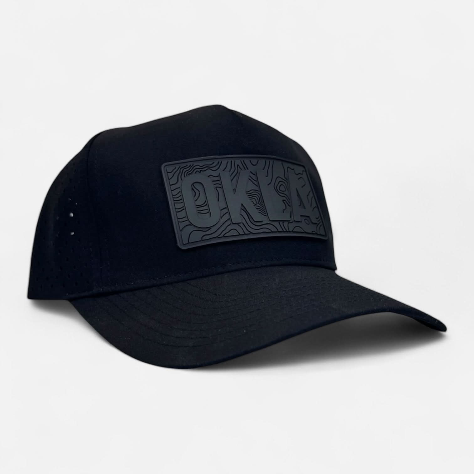 The OKLA Navigator Hat is a black baseball cap adorned with a vintage-style 3D "OKLA" patch on the front panel. It features a flat brim and small ventilation perforations, complete with sweat-wicking technology for comfort, all presented against a simple white background.