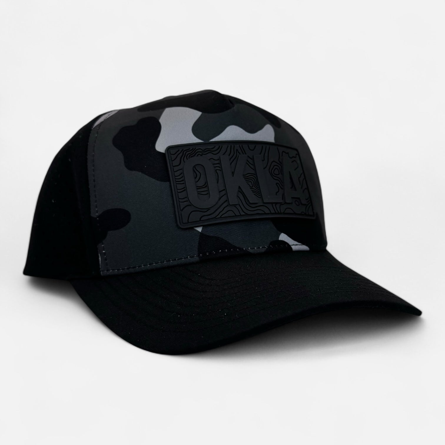 The OKLA Navigator Hat showcases a vintage-inspired 3D patch with the letters OKLA embossed on its black and gray camo front panel, complemented by a plain white background. This baseball cap is equipped with sweat-wicking technology to enhance comfort.