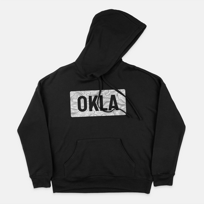 The OKLA Navigator BOLD Hoodie, made from recycled cotton for a comfortable unisex fit, is presented in black with a front pocket. The design includes intricate swirls and bold black letters spelling "OKLA" in a rectangular format. The hood is shown laid flat on a white background.