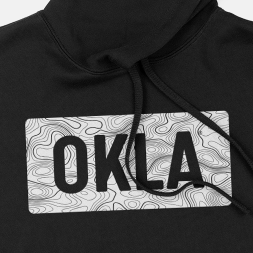 The OKLA Navigator BOLD Hoodie is a black hoodie showcasing a graphic design on the chest, which includes the word OKLA in bold, black letters set against a white background with a topographic map pattern. Crafted from recycled cotton, it provides a comfortable unisex fit and features adjustable drawstrings on the sides.