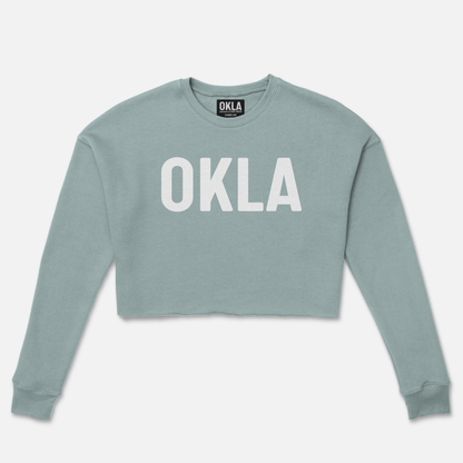 The OKLA Original Crop Sweatshirt is a long-sleeve, light blue fleece top featuring "OKLA" in bold white capital letters. Its simple yet trendy design with ribbed cuffs makes it perfect for making a fashion-forward statement.