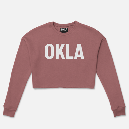 Introducing the OKLA Original Crop Sweatshirt: a chic mauve fleece design adorned with bold white "OKLA" lettering on the front. This fashionable piece, complete with long sleeves and a round neckline, is deserving of any fashion magazine spotlight.