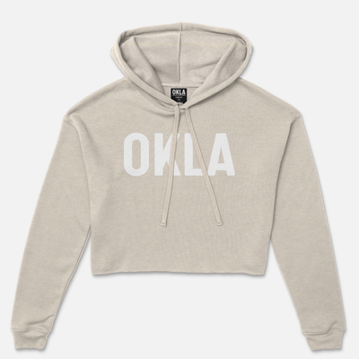 The OKLA Original Crop Hoodie, worthy of a fashion magazine, showcases long sleeves and a cozy fleece texture in a chic beige color. Its trendy design features a front pouch pocket and "OKLA" boldly printed in white across the chest, complemented by a functional drawstring hood.