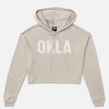 The OKLA Original Crop Hoodie, worthy of a fashion magazine, showcases long sleeves and a cozy fleece texture in a chic beige color. Its trendy design features a front pouch pocket and "OKLA" boldly printed in white across the chest, complemented by a functional drawstring hood.