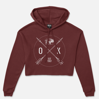 The OKLA Shield - Crop Hoodie, in maroon with a raw hem, showcases a graphic design featuring the letters O K and crossed arrows. It includes the words EST 1907 and an outline of a bison to complete the circular front design, while its soft fleece lining ensures cozy comfort.