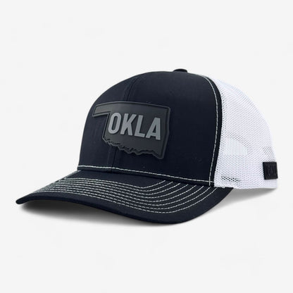 The OKLA Legacy Trucker Hat is a black baseball cap crafted with premium mesh fabric. It features a vintage-inspired PVC patch shaped like Oklahoma with the letters OKLA in white, and contrasting white stitching on the brim for additional detail.