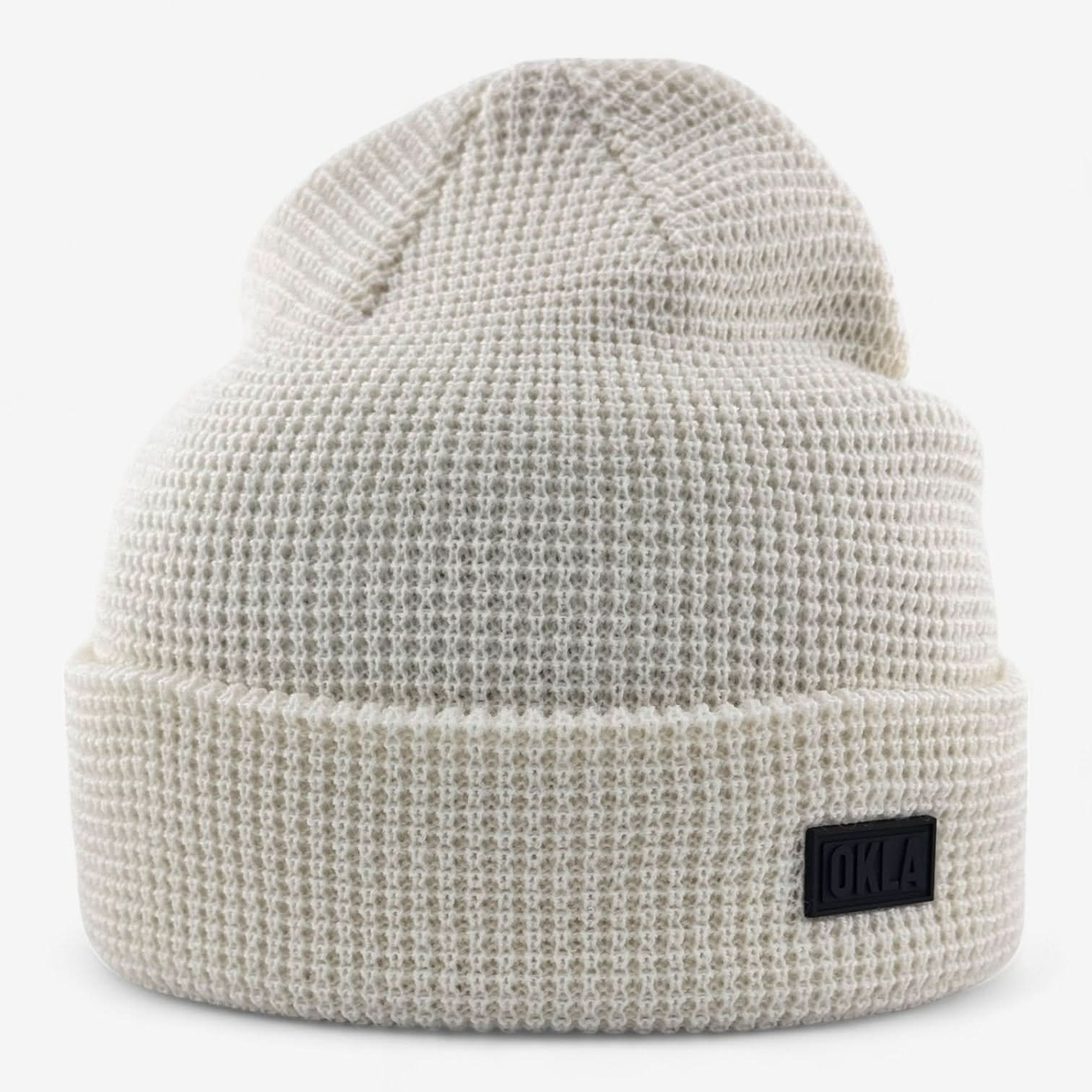 The OKLA Modern Beanie, in a beige waffle knit design with a textured pattern, is showcased against a plain background. It features a folded brim with a small black rectangular label adorned with subtle lettering and an OKLA rubber seal, highlighting its distinctive style.