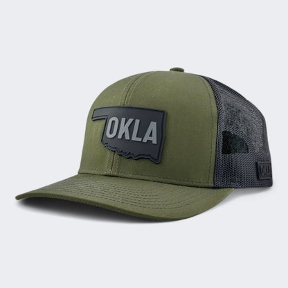 The OKLA Legacy Trucker Hat, in a military green shade, showcases a vintage-inspired PVC patch depicting the outline of Oklahoma with the text "OKLA." Made from premium mesh fabric, this hat combines style and comfort.