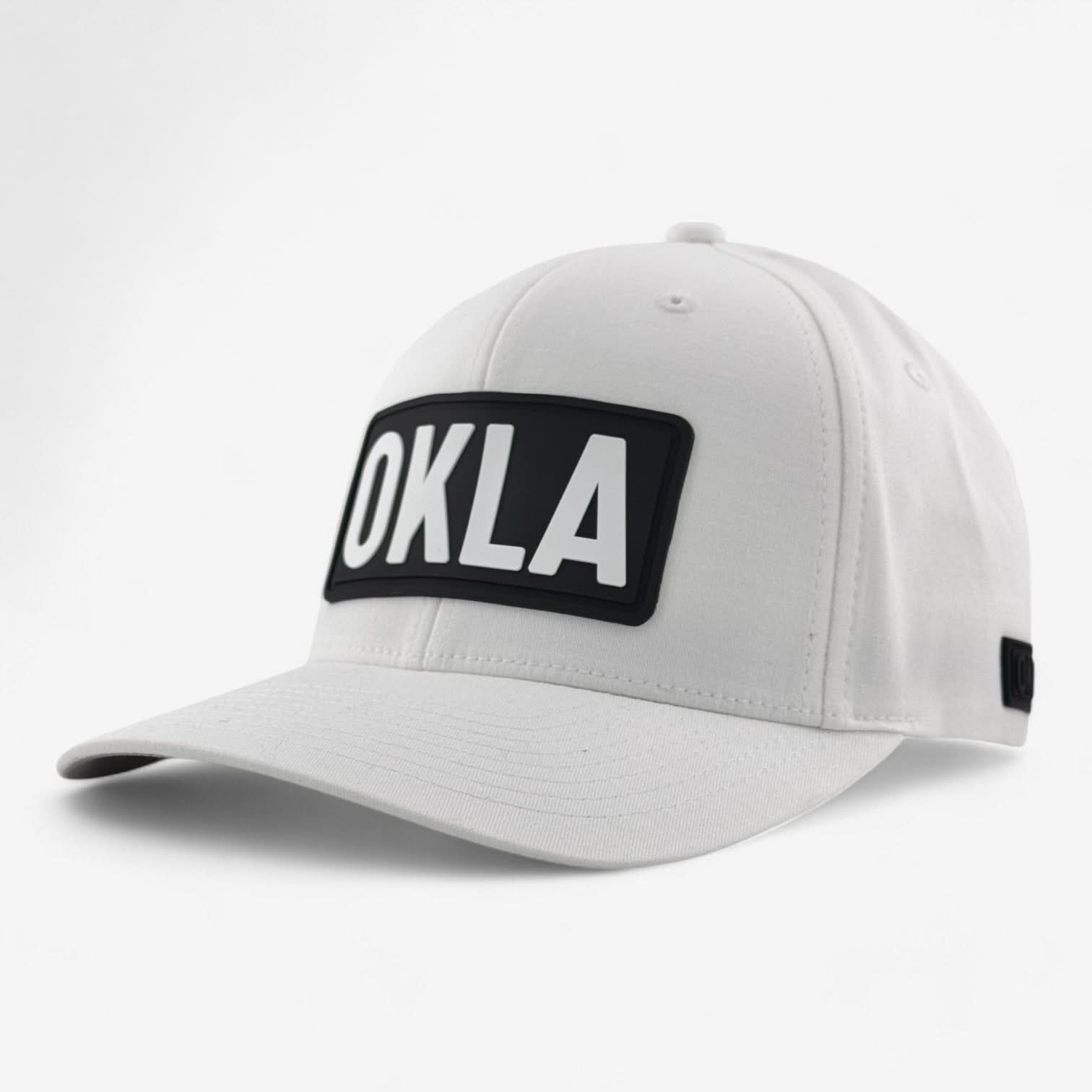 The OKLA Original Fitted Hat is a white baseball cap with a black rectangular patch displaying the letters OKLA in white. It features a curved visor, vent holes for breathability, and an elastic stretch band for added comfort.