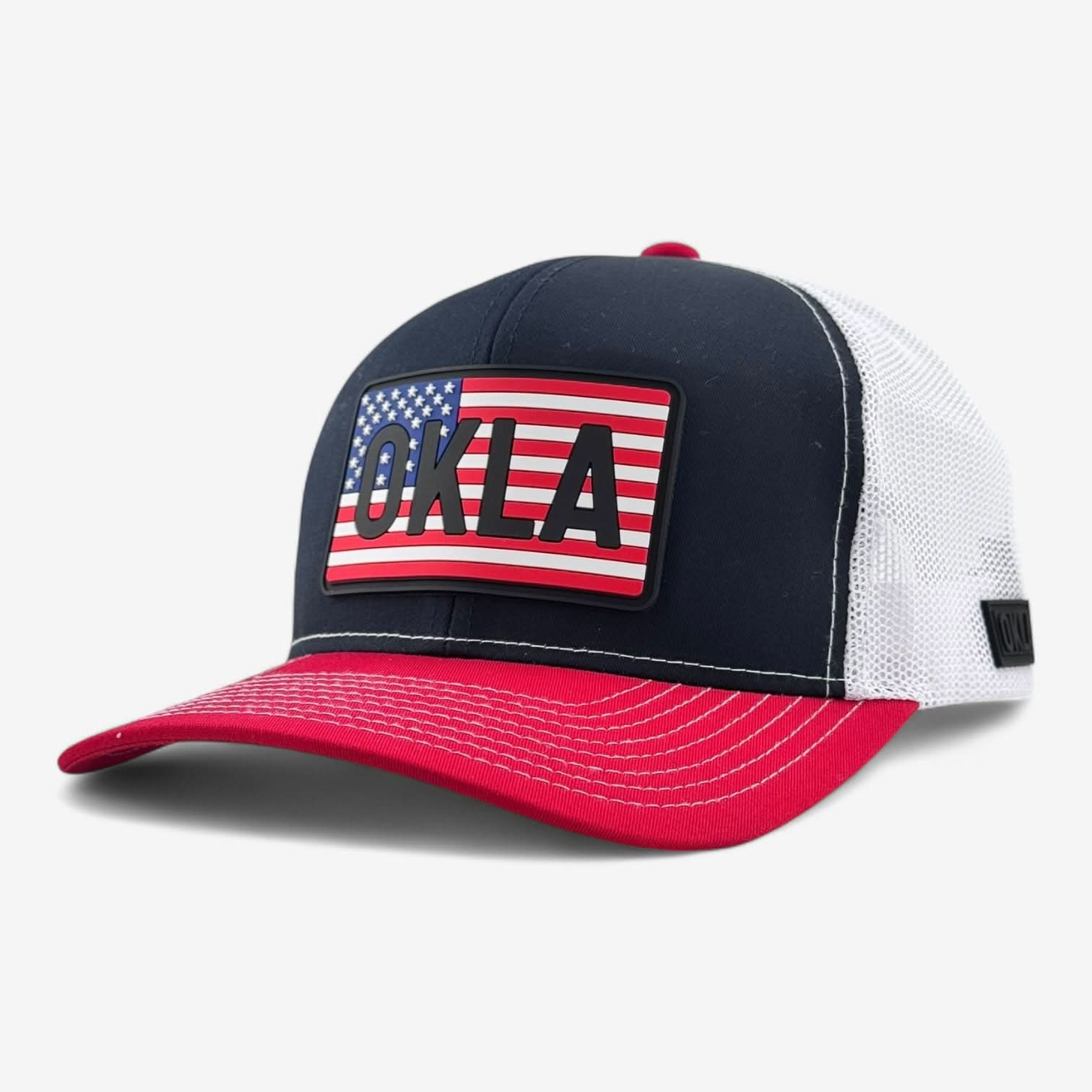 The OKLA Freedom Trucker Hat showcases a vintage-inspired 3D patch of the U.S. flag along with bold OKLA letters on its front. It sports a sophisticated black color and includes sweat-wicking technology, complemented by a white mesh back. The design is completed with a red bill accentuated by white stitching for a classic finish.