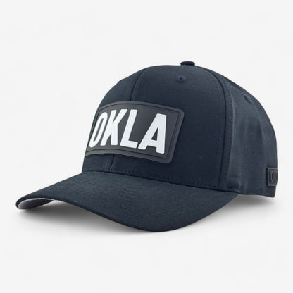 The OKLA Original Fitted Hat is a black baseball cap featuring a prominent, raised rectangular patch on the front with "OKLA" displayed in bold, white letters. This hat offers a comfortable fit with its elastic stretch band and curved visor. It comes with a curved brim and has an adjustable strap, all showcased against a plain white background.