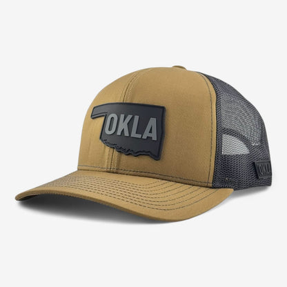 The OKLA Legacy Trucker Hat showcases a hunter brown design accompanied by a vintage-inspired PVC patch shaped like Oklahoma, prominently featuring "OKLA" on the front. Crafted with premium mesh fabric for breathability, it is the ideal accessory that melds style and comfort.