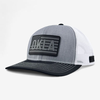 Introducing the OKLA Freedom Trucker Hat: a heather grey and white mesh baseball cap featuring a vintage-inspired 3D OKLA patch that resembles the American flag. The front panel is heather grey, with a black brim accented by white stitching, complemented by sweat-wicking technology in its white mesh back.