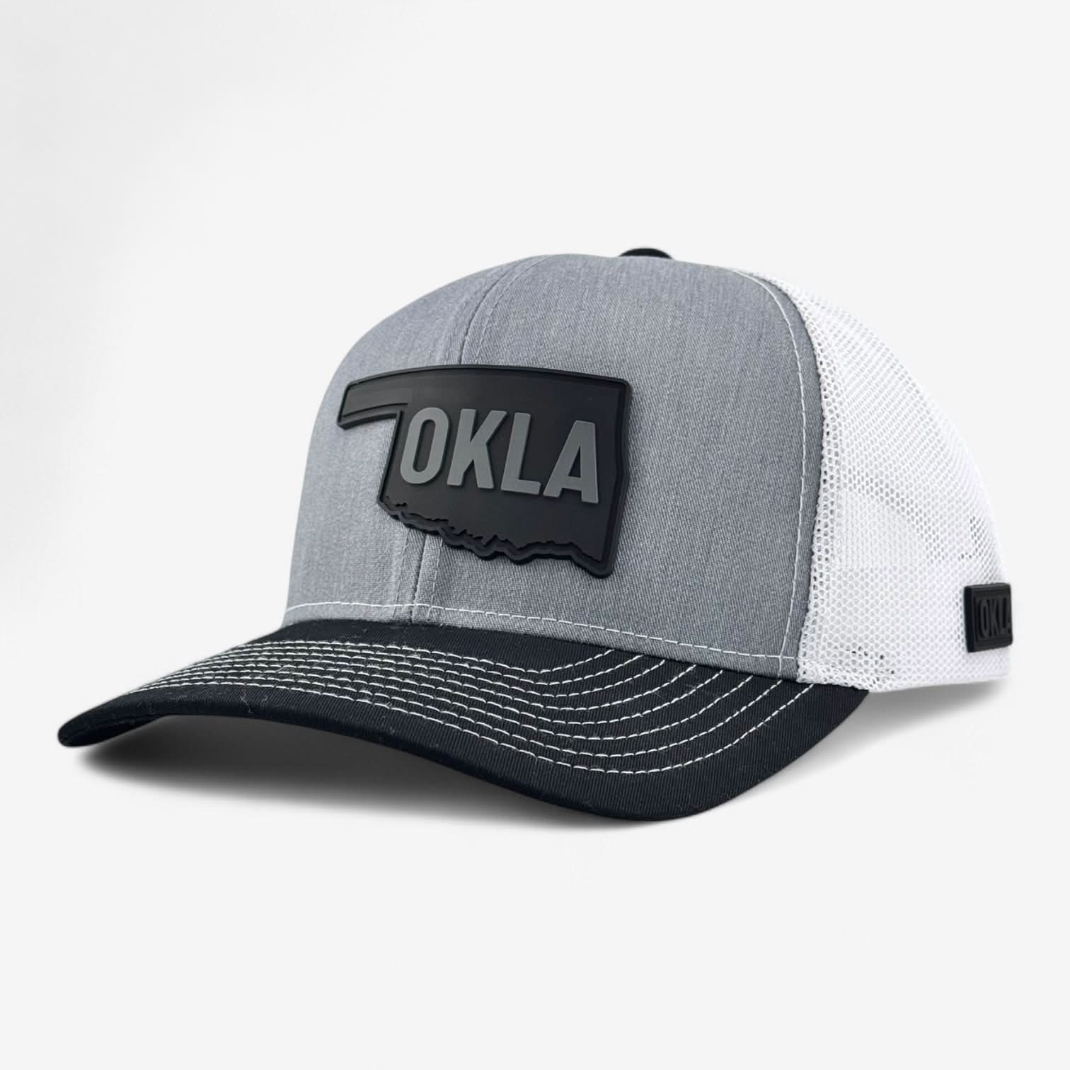 The OKLA Legacy Trucker Hat boasts a heather grey and white design with a vintage-inspired PVC patch shaped like Oklahoma on the front. It features a black bill for added style and premium mesh fabric that ensures comfort, along with a small matching patch on the side set against a plain white background.