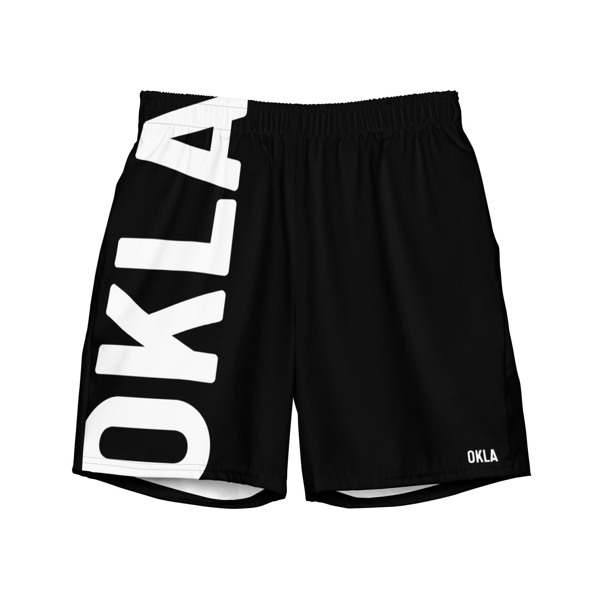 The OKLA Boardshorts are black athletic shorts displaying the word "OKLA" in large white letters vertically on the left leg and in smaller white letters on the bottom right. Made from sustainable fabric, these shorts offer a comfortable elastic waistband and quick-drying properties.