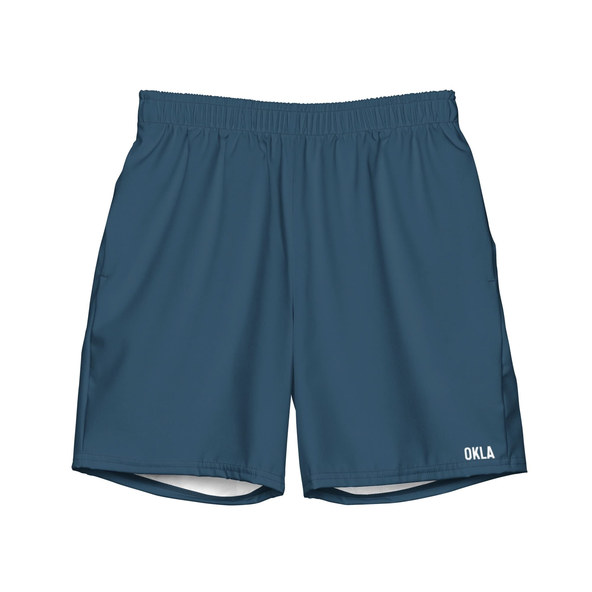 Introducing the OKLA Boardshorts: blue athletic shorts equipped with an elastic waistband and adorned with "OKLA" in white near the hem, specially designed to be quick-drying for your active lifestyle.