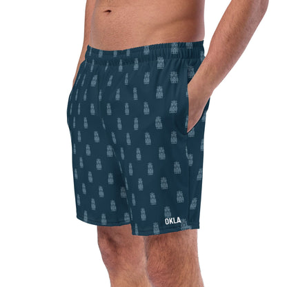 A person wears Tiki Boardshorts, showcasing a pattern of small gray rectangles on a dark blue background, with "OKLA" printed in white near the hem. With their quick-dry fabric and island-inspired design, these shorts are ideal for casual outings, featuring pockets that allow hands to rest comfortably.