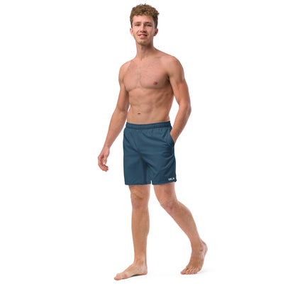 A man with curly hair stands barefoot, confidently posing in OKLA Boardshorts, which are blue in color. These quick-drying, water-repellent shorts enhance his shirtless look against the white background with their functional design.