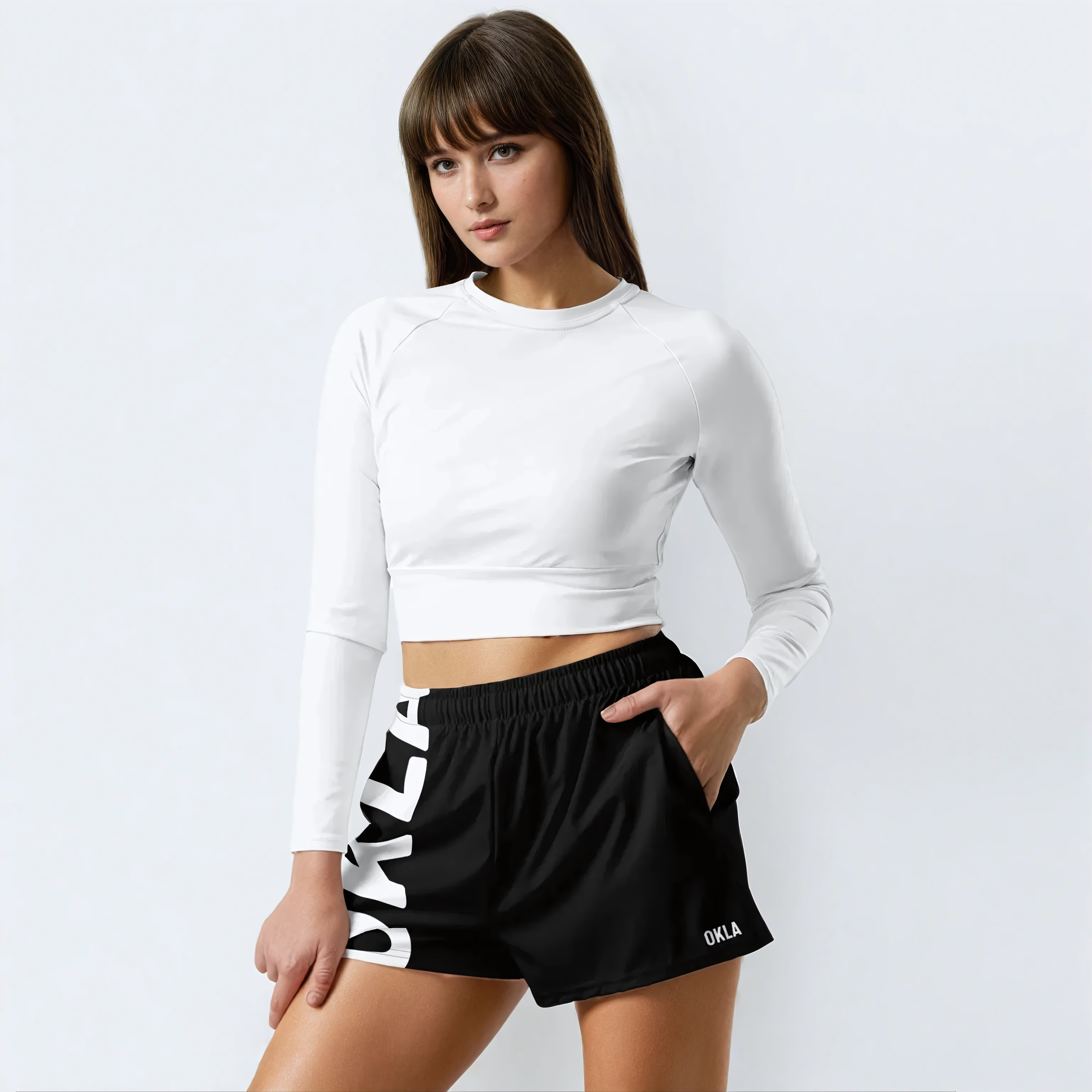 A woman stands against a plain white background wearing a white long-sleeve crop top and "The OKLA - Women’s Shorts," which are black athletic shorts featuring bold white lettering. These shorts, made from moisture-wicking fabric, provide both comfort and style, with one hand casually resting in her pocket.