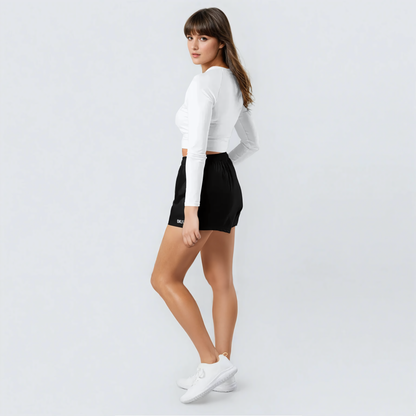 A woman poses against a plain white background, looking over her shoulder in a white long-sleeve moisture-wicking shirt, The OKLA - Women’s Shorts in black, and white sneakers.