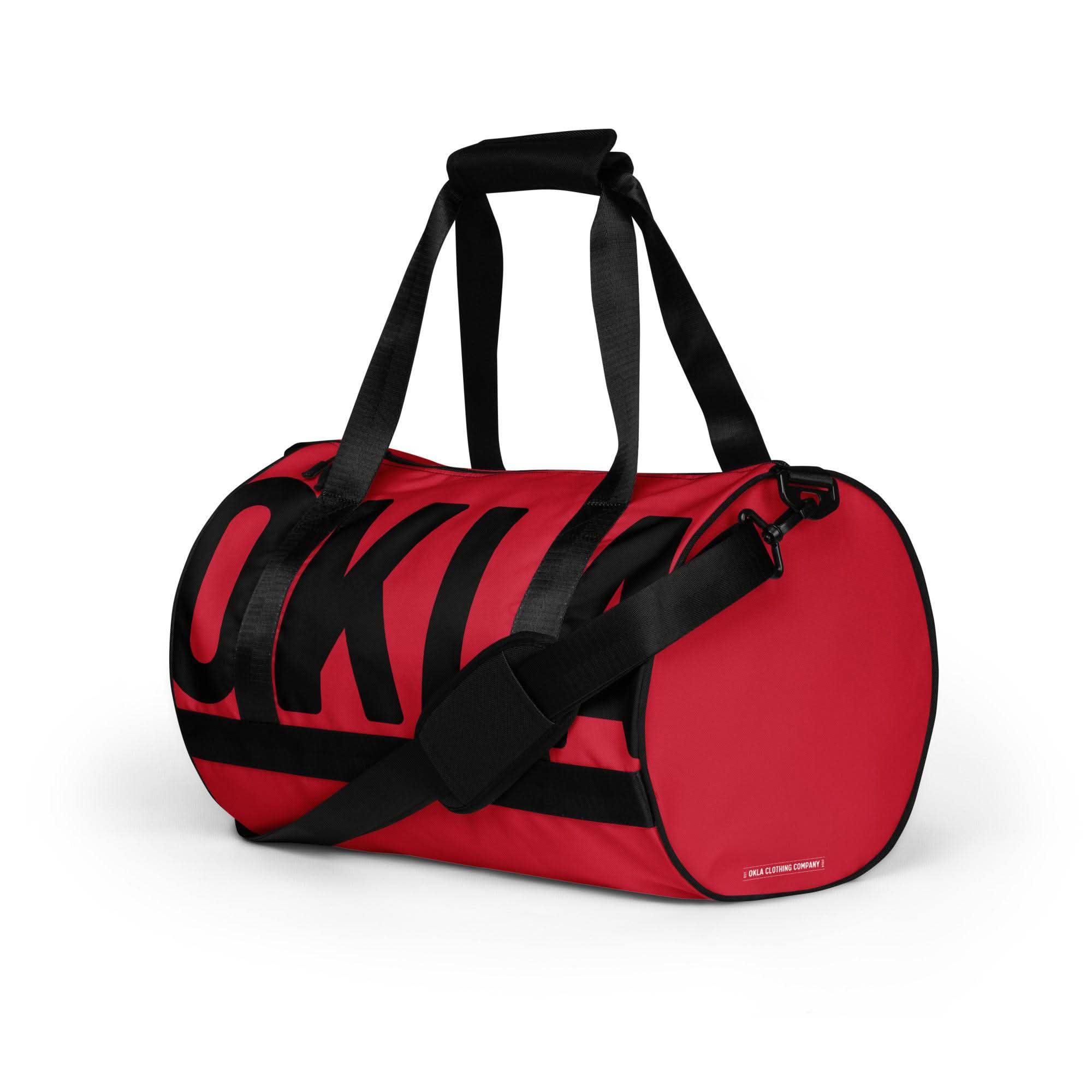 Introducing The OKLA - Gym Bag Red Series, a sporty and stylish design featuring a red cylindrical shape crafted from sturdy, water-resistant fabric. It boasts bold black lettering and sleek black handles. For added convenience, this gym bag includes an adjustable shoulder strap for easy carrying.