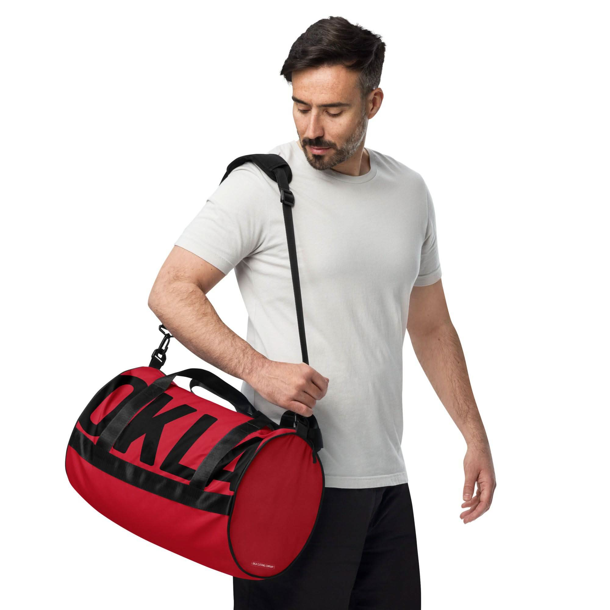 A man in a light gray t-shirt and black pants is carrying "The OKLA - Gym Bag Red Series" in red and black, with the letters OKLA partially visible. Made from sturdy, water-resistant fabric, he looks down at the bag while holding the shoulder strap against the plain white background.