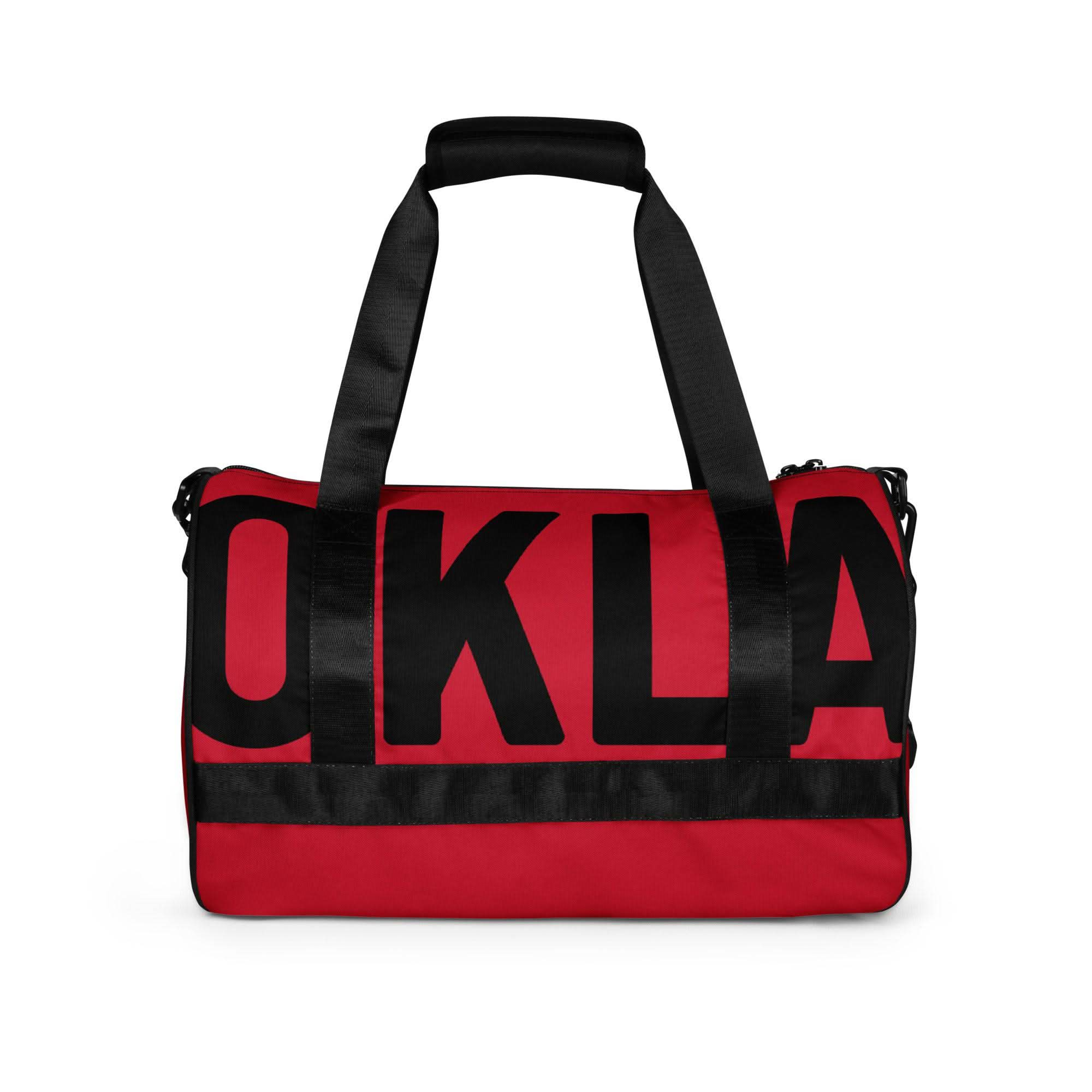 Introducing The OKLA - Gym Bag Red Series: This red and black gym bag features durable fabric and black handles. Its front is prominently adorned with large, bold black letters spelling "OKLA," making it an ideal choice for your active lifestyle.