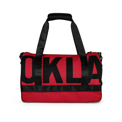 Introducing The OKLA - Gym Bag Red Series: This red and black gym bag features durable fabric and black handles. Its front is prominently adorned with large, bold black letters spelling "OKLA," making it an ideal choice for your active lifestyle.