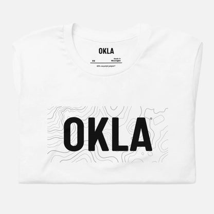 The OKLA Navigator Tee is a neatly folded white T-shirt adorned with the bold 'OKLA' logo atop a delicate, abstract contour line design. Made from 40% recycled polyester, it guarantees both comfort and adventure. The inner neck label states "OKLA, Made in Melbourne," with the size indicated as XS.