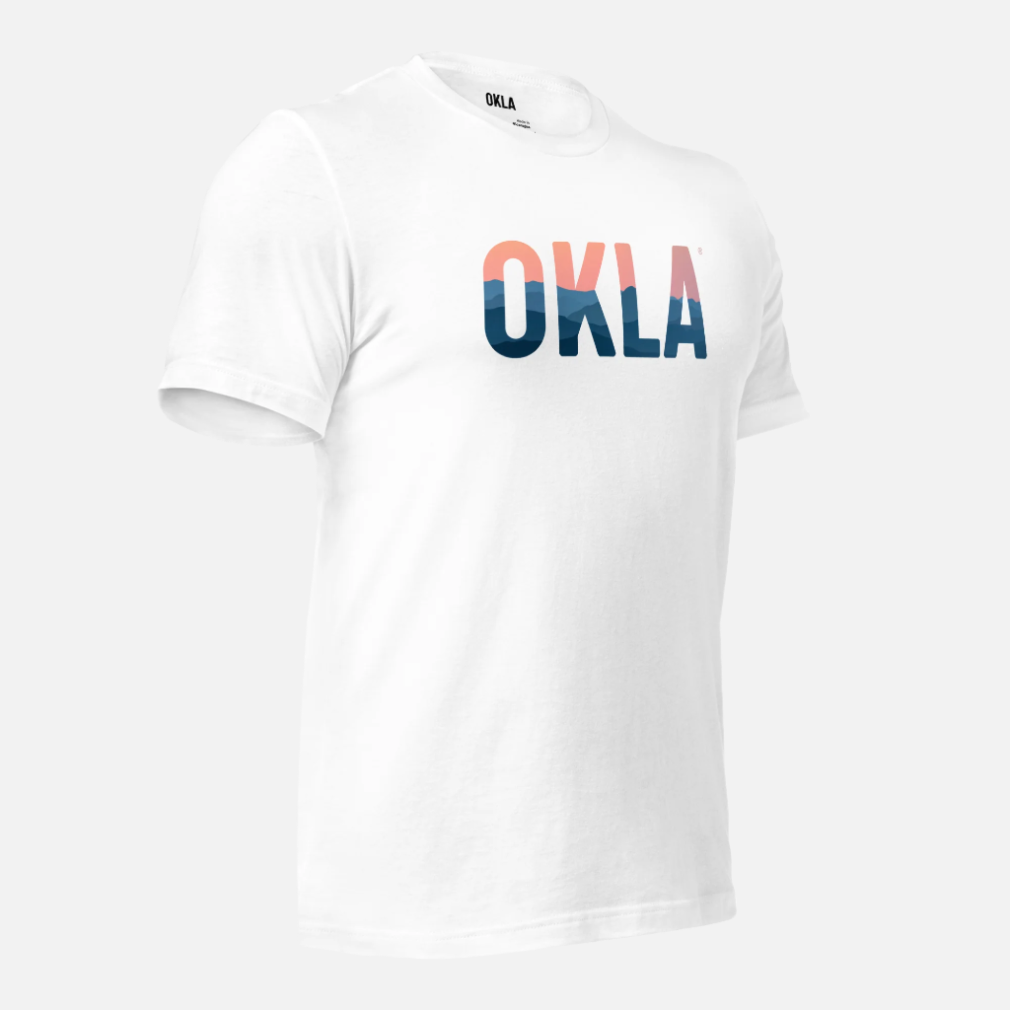 The OKLA Horizon Tee features a striking gradient design in black, pink, and blue on the front. Made from recycled cotton, this white T-shirt seamlessly blends adventurous comfort with style. Photographed against a plain background, it's an ideal choice for those looking to make a bold statement in sustainable fashion.